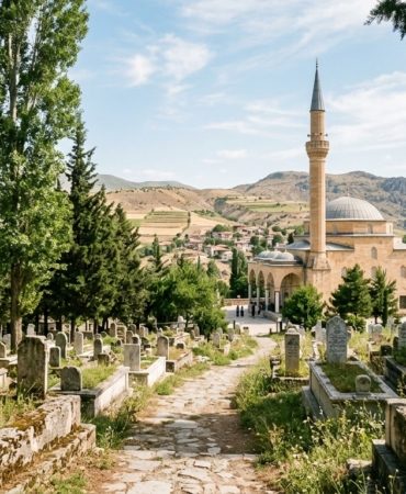 A cinematic, wide-angle shot of a serene Anatolian landscape at noon, featuring a traditional stone mosque with a single slender minaret in the background. In the foreground, a peaceful cemetery is nestled among tall, dark green cypress trees and weathered marble headstones. The warm, golden sunlight of a summer afternoon filters through the branches, casting soft shadows across the quiet grounds. The atmosphere is one of profound peace, dignity, and timeless remembrance, captured in a high-quality, editorial photography style with a soft, respectful color palette.
