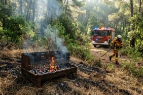 A cinematic, editorial-style photograph capturing a dramatic scene in a Mediterranean pine forest. In the foreground, an abandoned metal barbecue grill sits on scorched dry grass, with bright orange embers still glowing and thin trails of smoke rising into the air. The background features a lush, green forest under a hazy, sunlight-dappled canopy. In the middle ground, a fire engine with its emergency lights flashing is parked on a nearby roadside, and a firefighter in protective gear is seen rushing toward the smoldering fire with a water hose. The lighting is intense and atmospheric, emphasizing the contrast between the small spark of fire and the vast, fragile beauty of the surrounding nature.