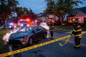 A cinematic, wide-angle editorial photograph of a single-vehicle accident on a quiet residential street at dusk. A modern sedan with a crumpled front bumper sits stationary, with a visible plume of white gas or vapor escaping from under the hood, illuminated by the strobing red and blue lights of emergency vehicles. In the background, the blurred lights of a fire truck and a police car create a sense of urgency. Firefighters in reflective protective gear are seen in the mid-ground, securing the area with bright yellow caution tape. The atmosphere is tense but controlled, capturing the moment a potential disaster is averted in a peaceful neighborhood setting.